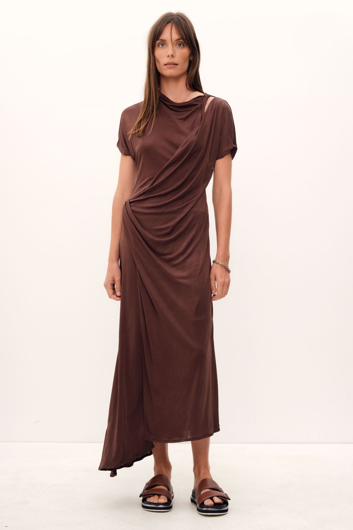 The Aphrodite Draped Dress, exquisitely crafted in silk viscose jersey of a coco hue, boasts a relaxed silhouette with an asymmetric hem and sleeves, a cinched waistline that gracefully drapes, a delicate stretch fabric, and a midi length with a sublime twist detail across the neck and shoulder.