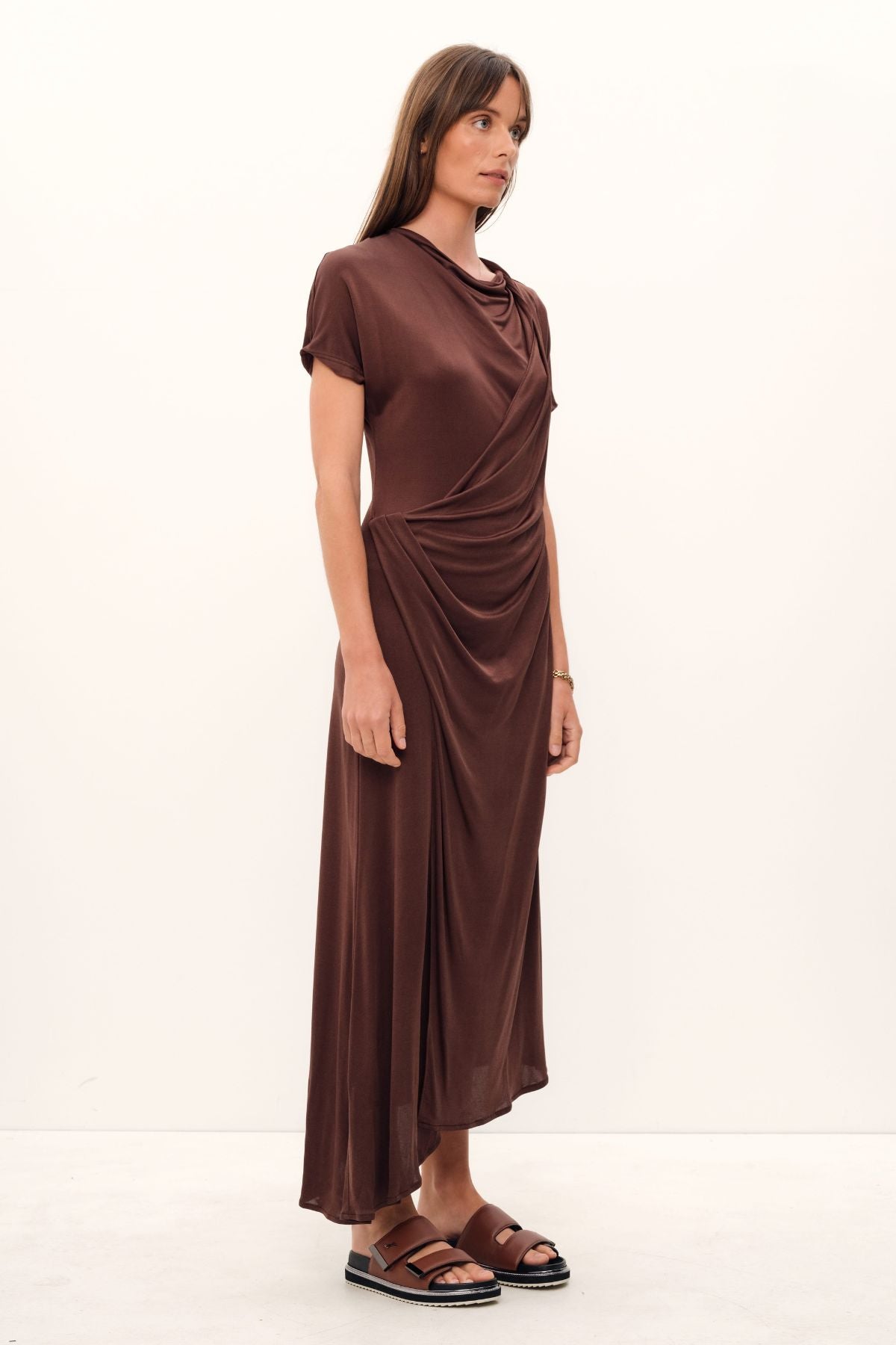 The Aphrodite Draped Dress, exquisitely crafted in silk viscose jersey of a coco hue, boasts a relaxed silhouette with an asymmetric hem and sleeves, a cinched waistline that gracefully drapes, a delicate stretch fabric, and a midi length with a sublime twist detail across the neck and shoulder.
