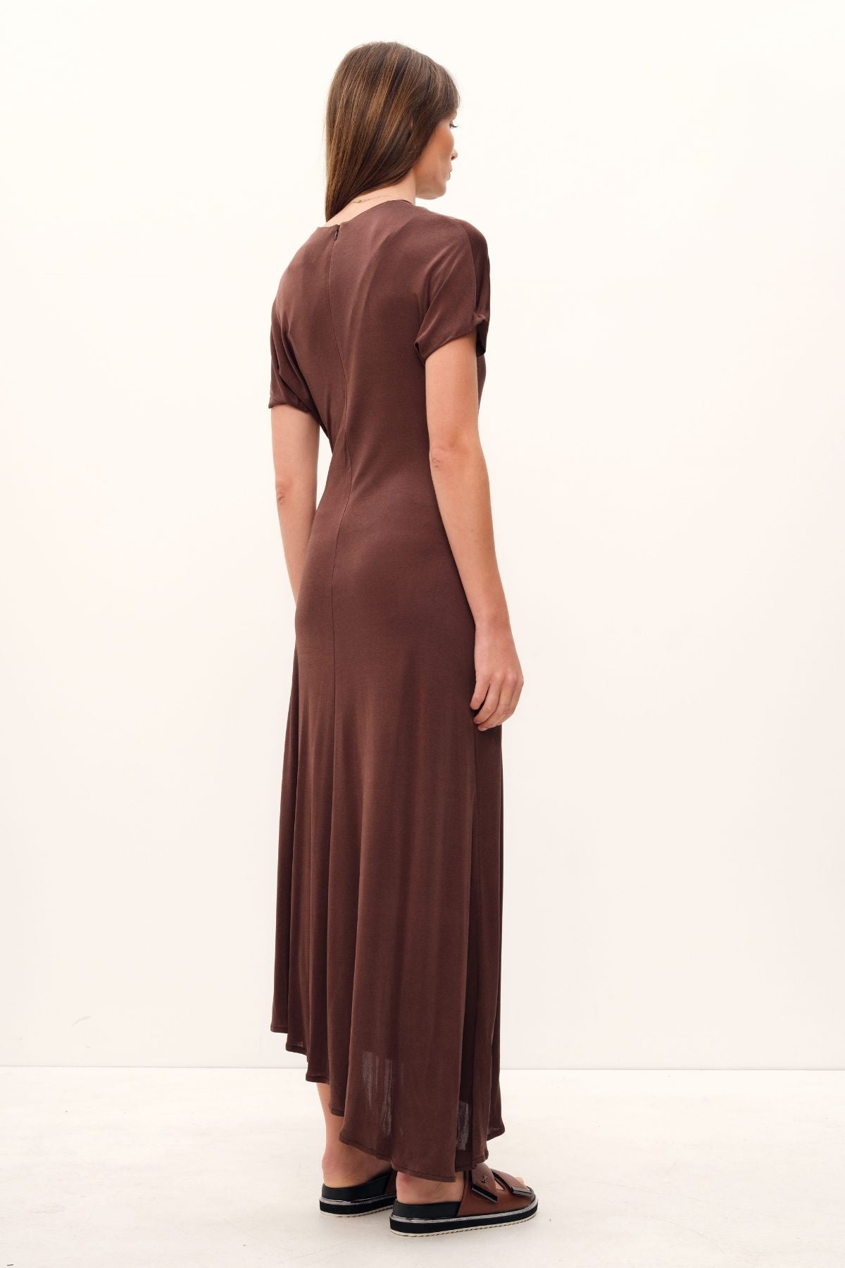 The Aphrodite Draped Dress, exquisitely crafted in silk viscose jersey of a coco hue, boasts a relaxed silhouette with an asymmetric hem and sleeves, a cinched waistline that gracefully drapes, a delicate stretch fabric, and a midi length with a sublime twist detail across the neck and shoulder.