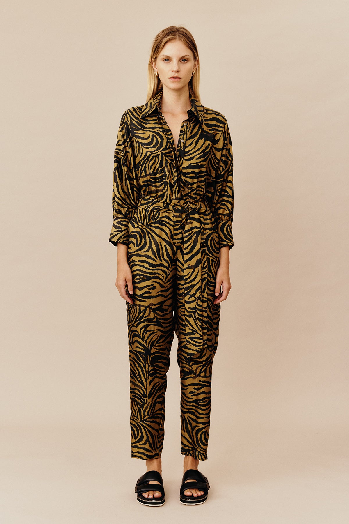 Model wearing Australian women’s fashion designer GINGER & SMARTS’ Feline Jumpsuit, crafted from a silk twill in olive green and gold animal print with a tapered leg silhouette, shirt collar, button through front, half sleeve and elasticised waist with tie. 