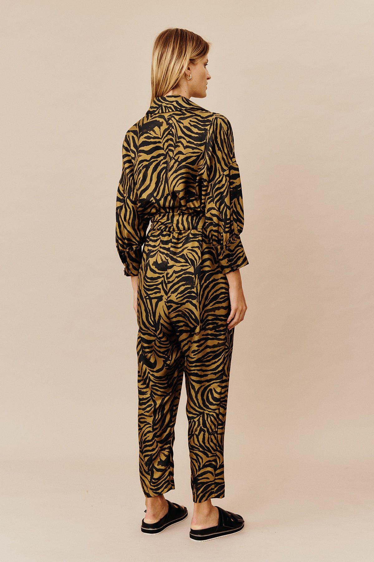 Back view of model wearing Australian women’s fashion designer GINGER & SMARTS’ Feline Jumpsuit, crafted from a silk twill in olive green and gold animal print with a tapered leg silhouette, shirt collar, button through front, half sleeve and elasticised waist with tie. 