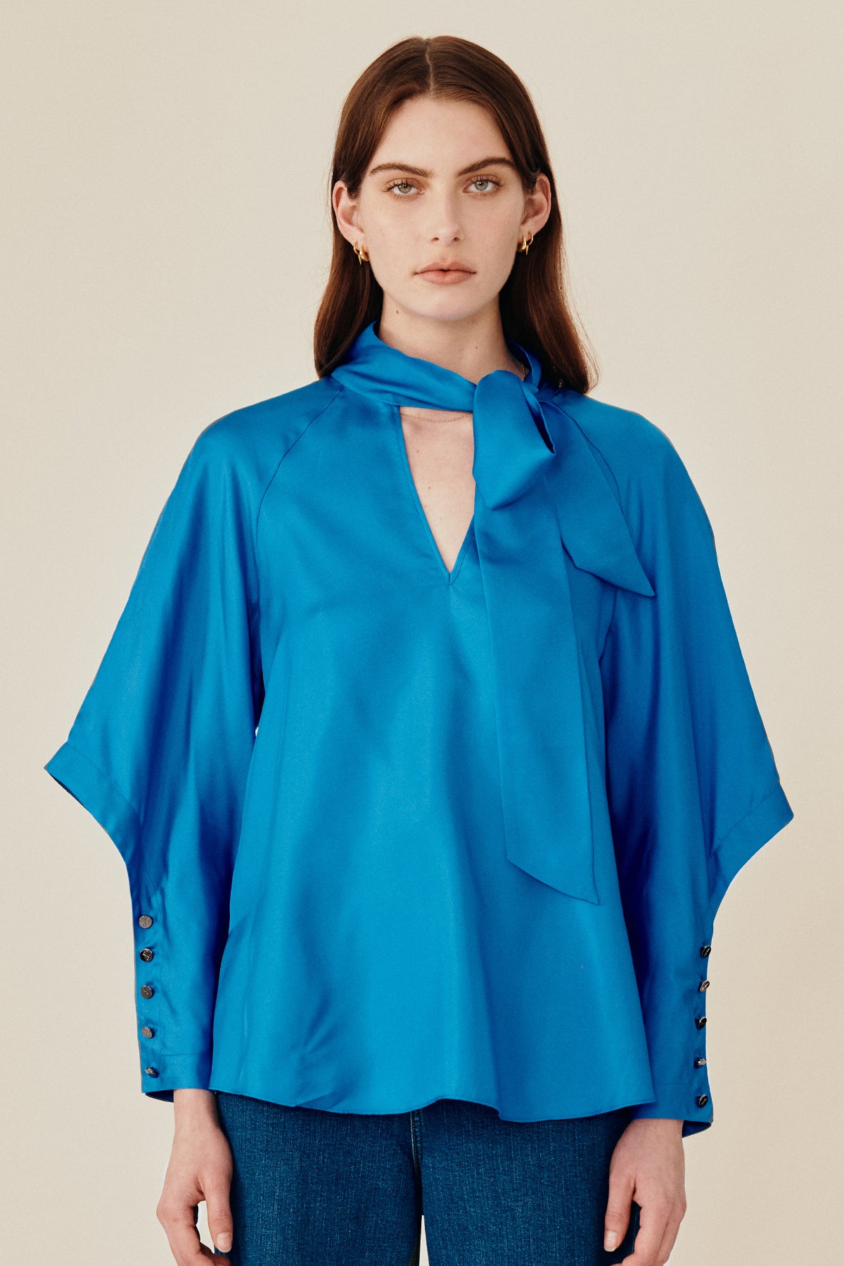 Model wearing Australian fashion designer GINGER & SMARTS' Libertine Blouse in blue silk in a silhouette that features a chic pussy necktie with a V-neck and angular sleeve with an extended cuff with a row of buttons. 
