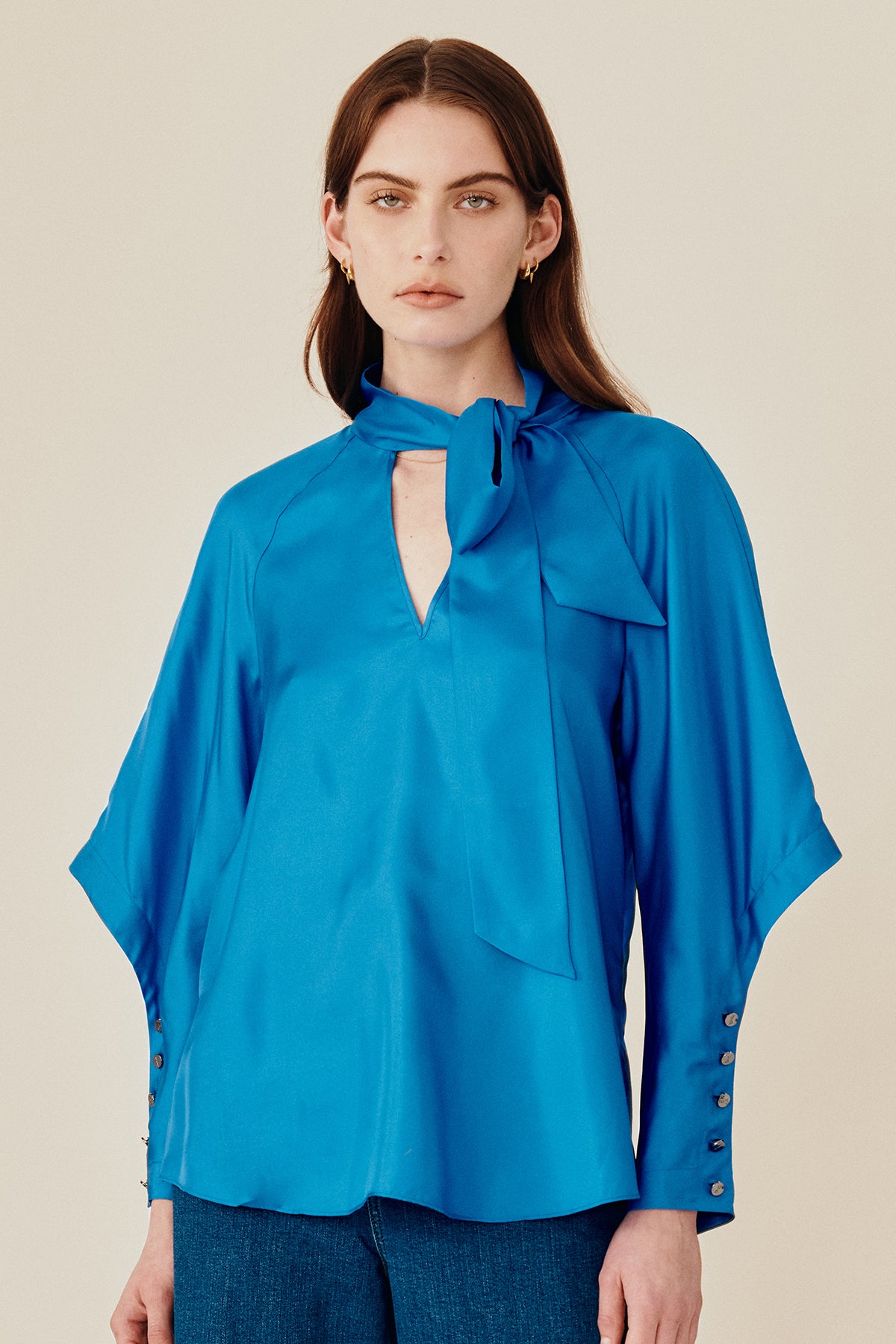 Model wearing Australian fashion designer GINGER & SMARTS' Libertine Blouse in blue silk in a silhouette that features a chic pussy necktie with a V-neck and angular sleeve with an extended cuff with a row of buttons. 