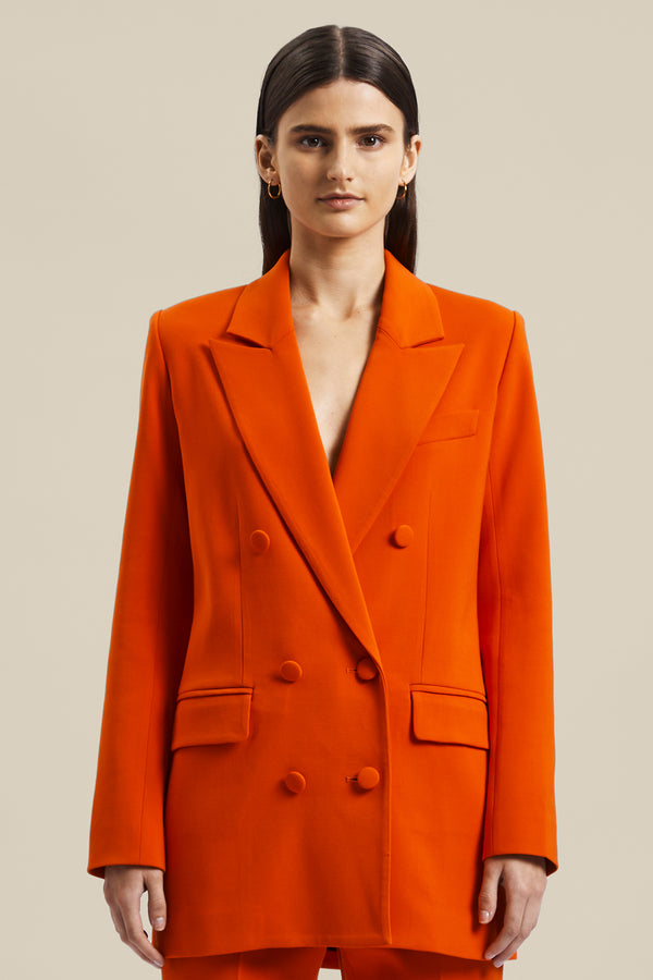 Model wearing the orange women’s Rhetoric Jacket from the Australian luxury fashion designer GINGER & SMART, featuring sharp lapel, long line blazer silhouette, pocket flaps and double breasted. Worn with the matching orange Rhetoric Pants. 