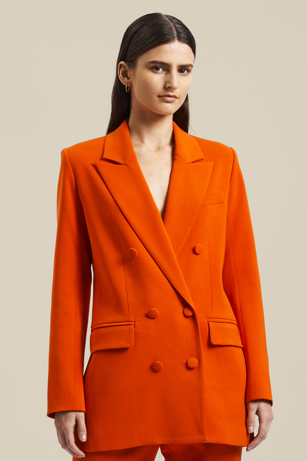 Model wearing the orange women’s Rhetoric Jacket from the Australian luxury fashion designer GINGER & SMART, featuring sharp lapel, long line blazer silhouette, pocket flaps and double breasted. Worn with the matching orange Rhetoric Pants. 