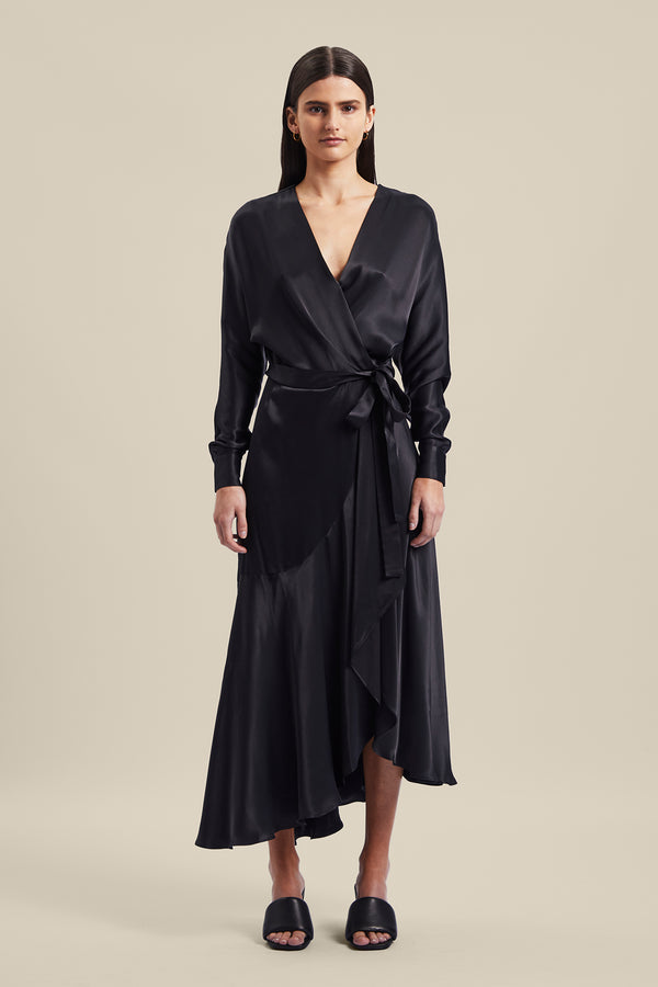 Model wearing black Nocturnal Wrap Dress from Australian luxury designer GINGER & SMART, featuring , long sleeves with wrist cuffs, waist tie, V neckline, midi length with bias cut asymmetrical hemline.