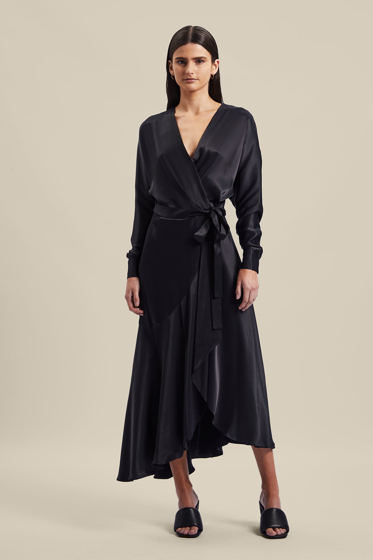 Model wearing black Nocturnal Wrap Dress from Australian luxury designer GINGER & SMART, featuring , long sleeves with wrist cuffs, waist tie, V neckline, midi length with bias cut asymmetrical hemline.