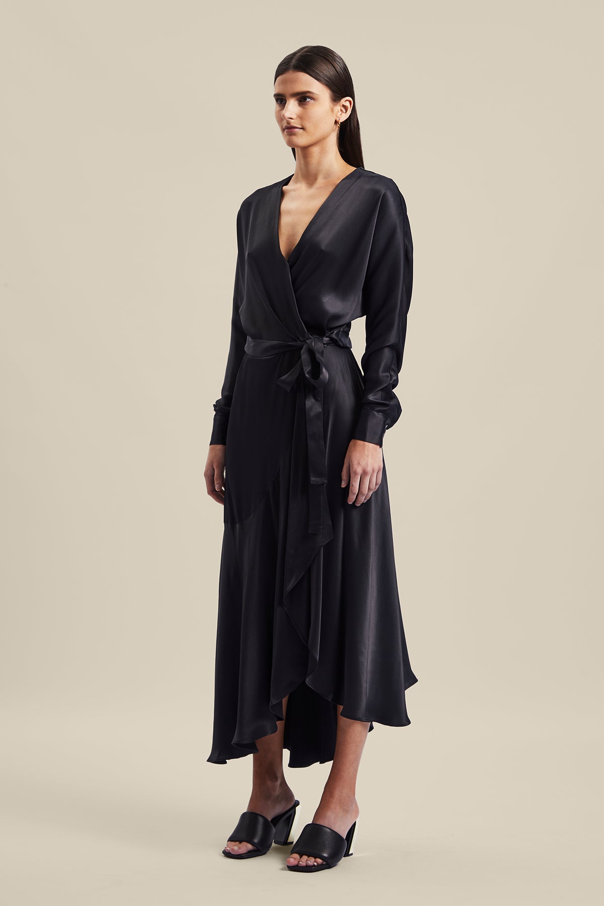 Model wearing black Nocturnal Wrap Dress from Australian luxury designer GINGER & SMART, featuring , long sleeves with wrist cuffs, waist tie, V neckline, midi length with bias cut asymmetrical hemline.