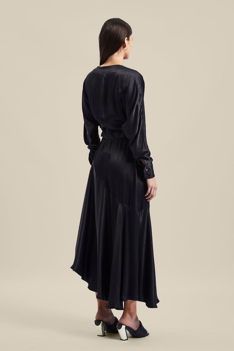 Back view of model wearing black Nocturnal Wrap Dress from Australian luxury designer GINGER & SMART, featuring , long sleeves with wrist cuffs, waist tie, V neckline, midi length with bias cut asymmetrical hemline.
